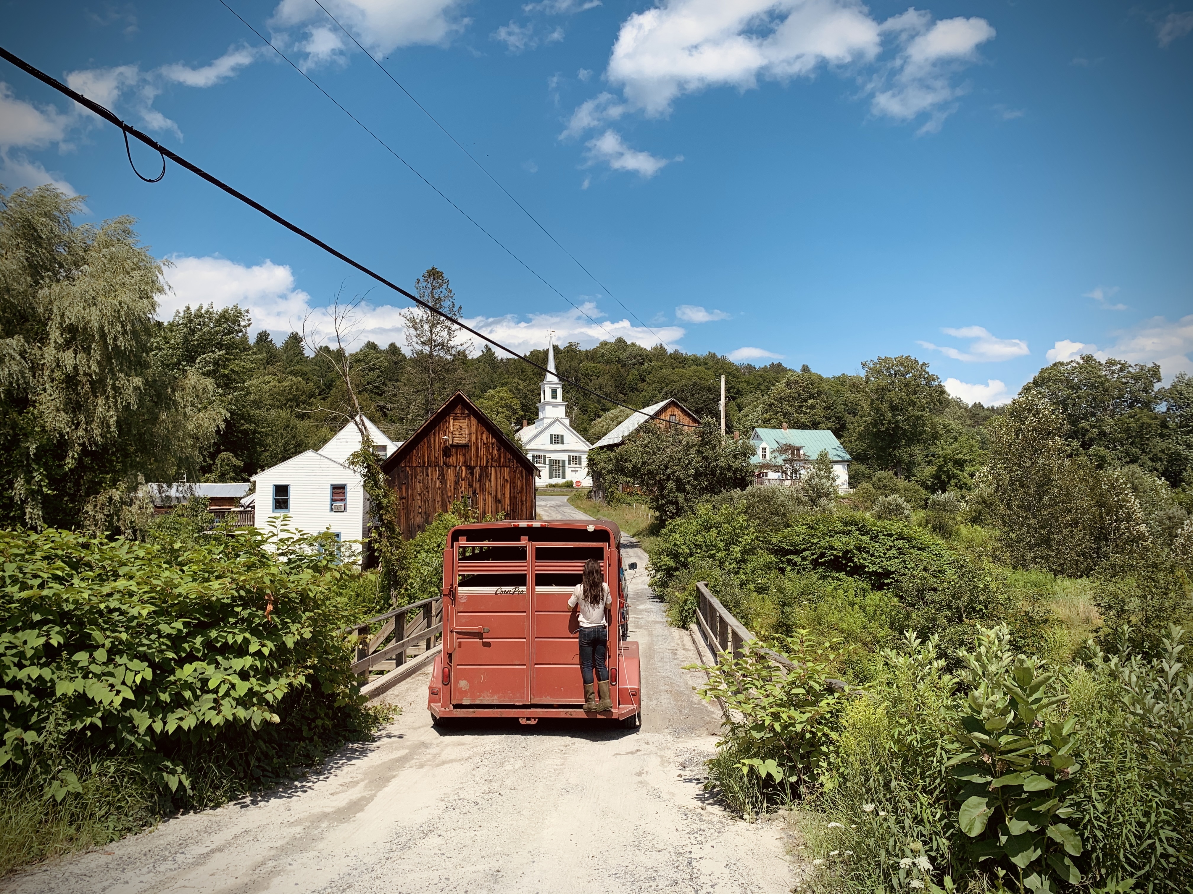 A woman rides the back of a red stock trailer over a dirt-road bridge into a Vermont village, white church steeple and barns ahead