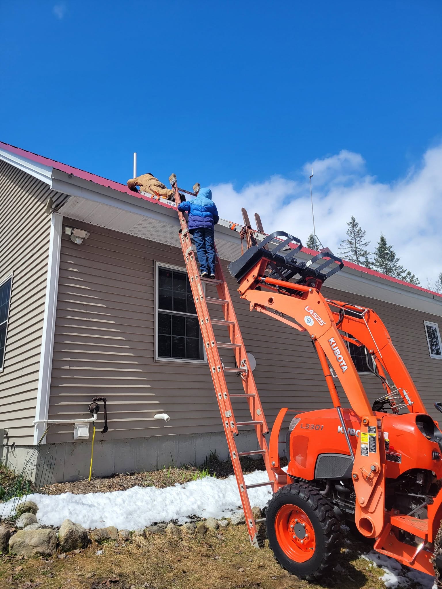 Two people work on a red metal roof from an orange extension ladder braced against a house, a compact tractor with a loader parked below on the last of the spring snow